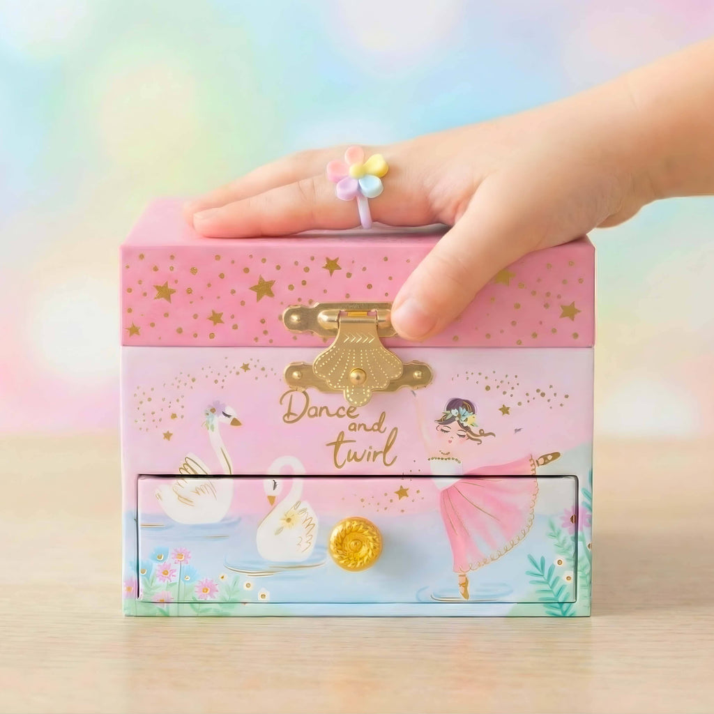 Ballerina front view with gold clam hardware with the phrase dance and twirl. Also on the box itself there are white swans alongside a ballerina in a white leotard and a pink skirt. There is also one drawer for storage  that has a blue lake and pink, blue, and white flowers. The top opens has gold stars and swan lake music will play.